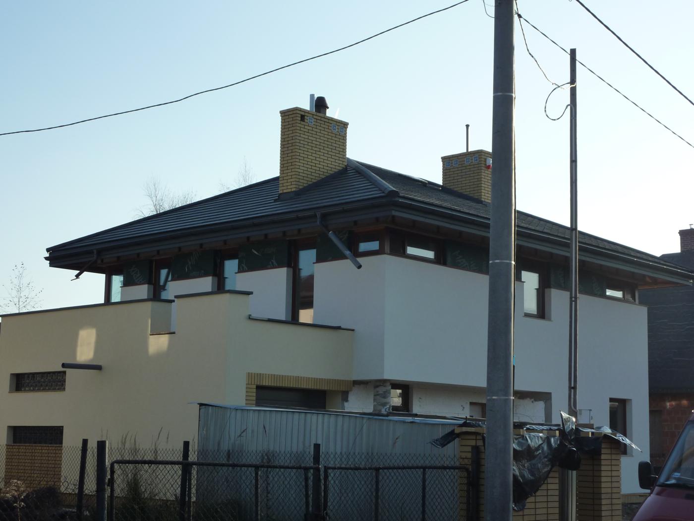 Nowoczesny dom w trakcie budowy z ciemną dachówką, jasną elewacją i ceglanymi kominami, widoczne elementy rusztowania i materiały budowlane.